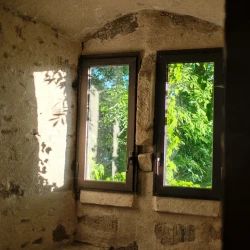 Alcôve avec des bancs en pierre de chaque côté, 2 fenêtres étroites séparées par un montant en pierre. Des branches d'arbre sont visibles et les rayons du soleil traversent et font des ombres sur le mur de gauche.