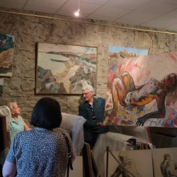 Dans une salle remplie de tableaux aux murs, 2 personnes en tiennent un qui représente un personnage masculin couché, habillé d'un seul slip. D'autres personnes regardent le tableau.