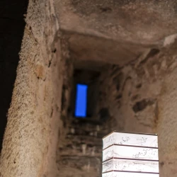 Une petite lampe avec un abat-jour en papier peint à l'aérographe éclaire en jaune, devant une meurtrière qu'on distingue avec la lueur bleu de fin du jour dans un renfoncement d'un mur très épais en pierre.