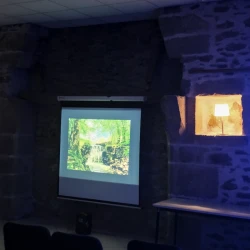 Dans une pièce aux murs en pierre plongée dans l'obscurité, un écran où l'on voit une cascade en photo. De chaque côté, des lumières dans une alcôve, au 1er plan à droite, une lueur bleue forte.