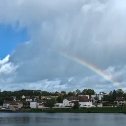 Vue d'un village avec une surface d'eau devant. Dans le ciel, des nuages, un peu de bleu et un arc-en-ciel partiel.