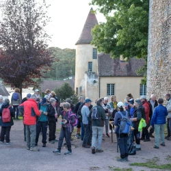 Un groupe de personnes rassemblées en train de se restaurer et boire, devant un édifice avec une tour, 1 arbre. À droite, on distingue un mur en pierre d'un bâtiment ancien.