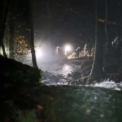 De nuit au premier plan : de l'eau qui coule en cascade, à l'arrière plan des marcheurs équipés de frontales passent en contre-bas sur un pont qui enjambe une rivière.