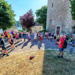 Un groupe de marcheurs sur une esplanade en semi ombre. À droite une vieille tour en pierre, au centre des arbres, et plus loin un autre bâtiment ancien, des maisons, et un lac et une forêt en arrière plan.