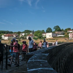 Un groupe de marcheurs sur un barrage : la partie la plus proche est à l'ombre, le reste au soleil. Au loin on aperçoit un village.