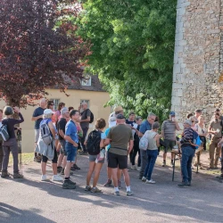 Un groupe de personnes autour d'une table qui boivent un verre et grignotent au pied d'une tour en vieilles pierres. En arrière plan de la végétation et un autre bâtiment ancien. Sous le soleil.